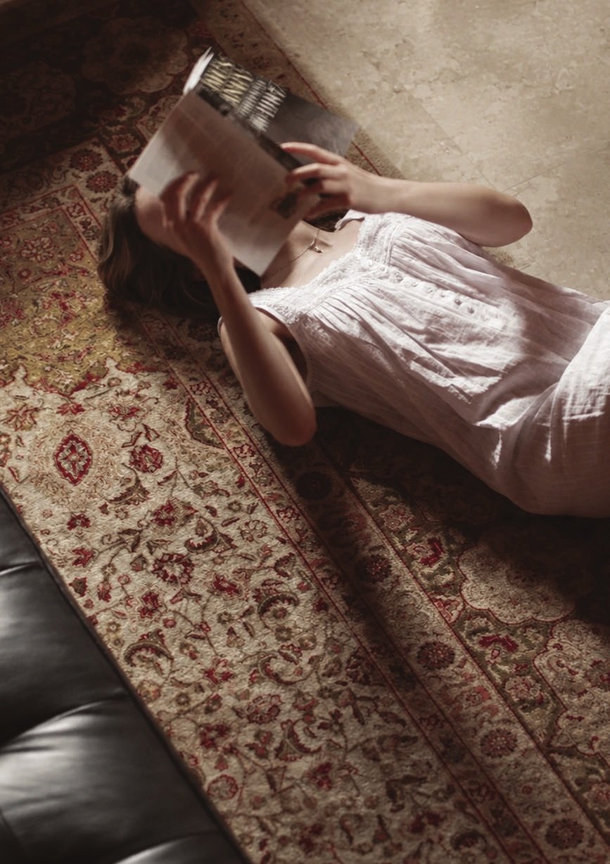 A woman lying on a patterned rug, reading a book held above her face